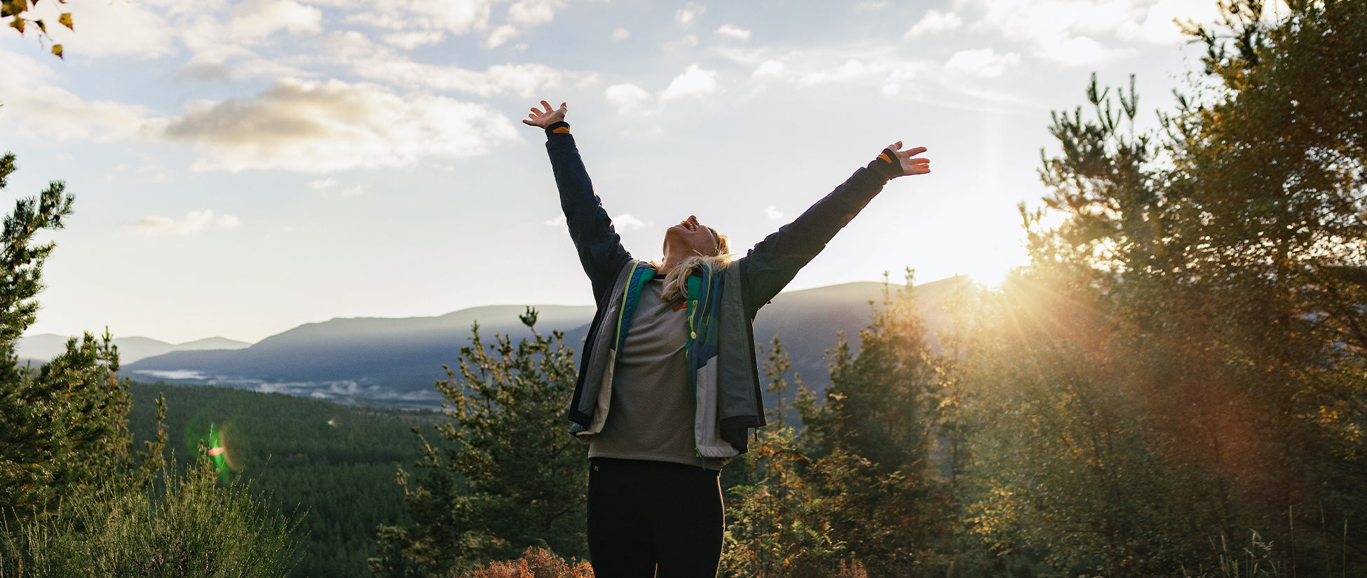 A woman, arms outstretched atop a wooded valley, golden light