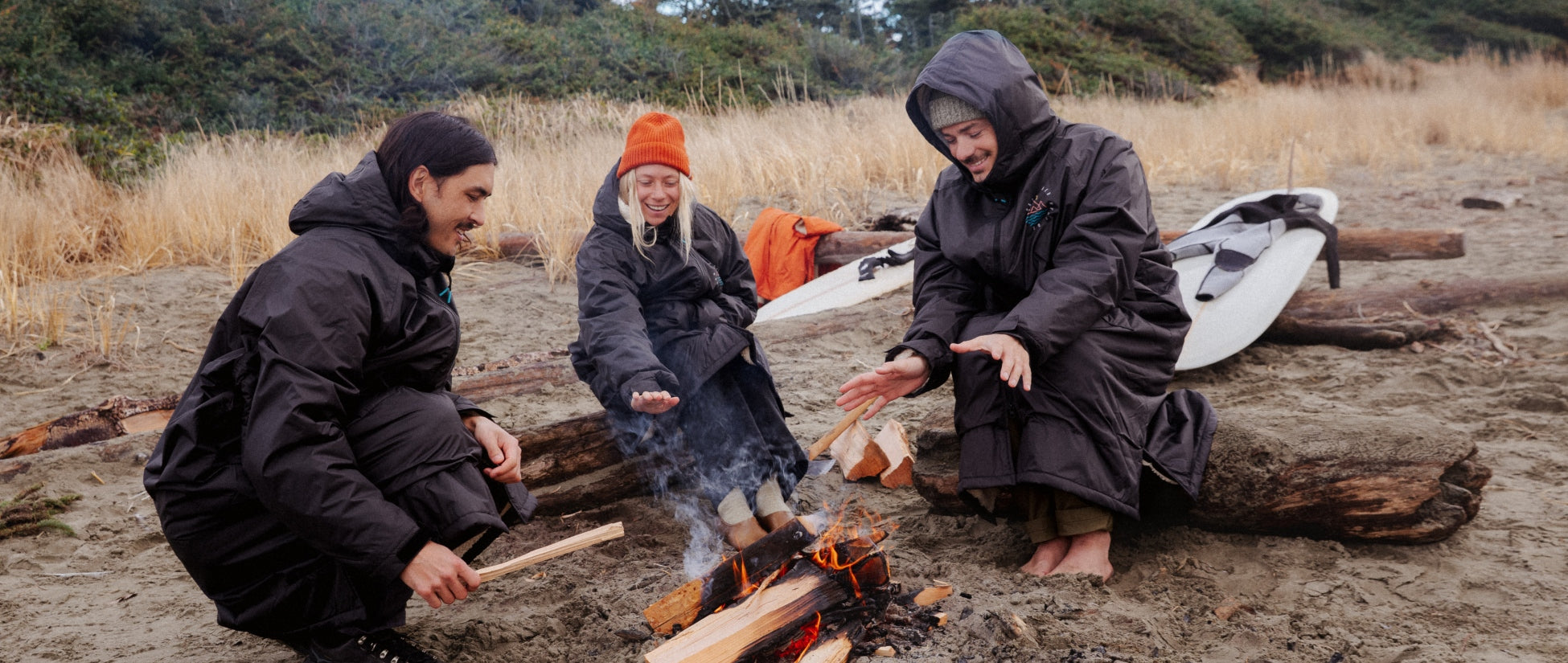 Three people on the beach huddled round a campfire, wearing black changing robes